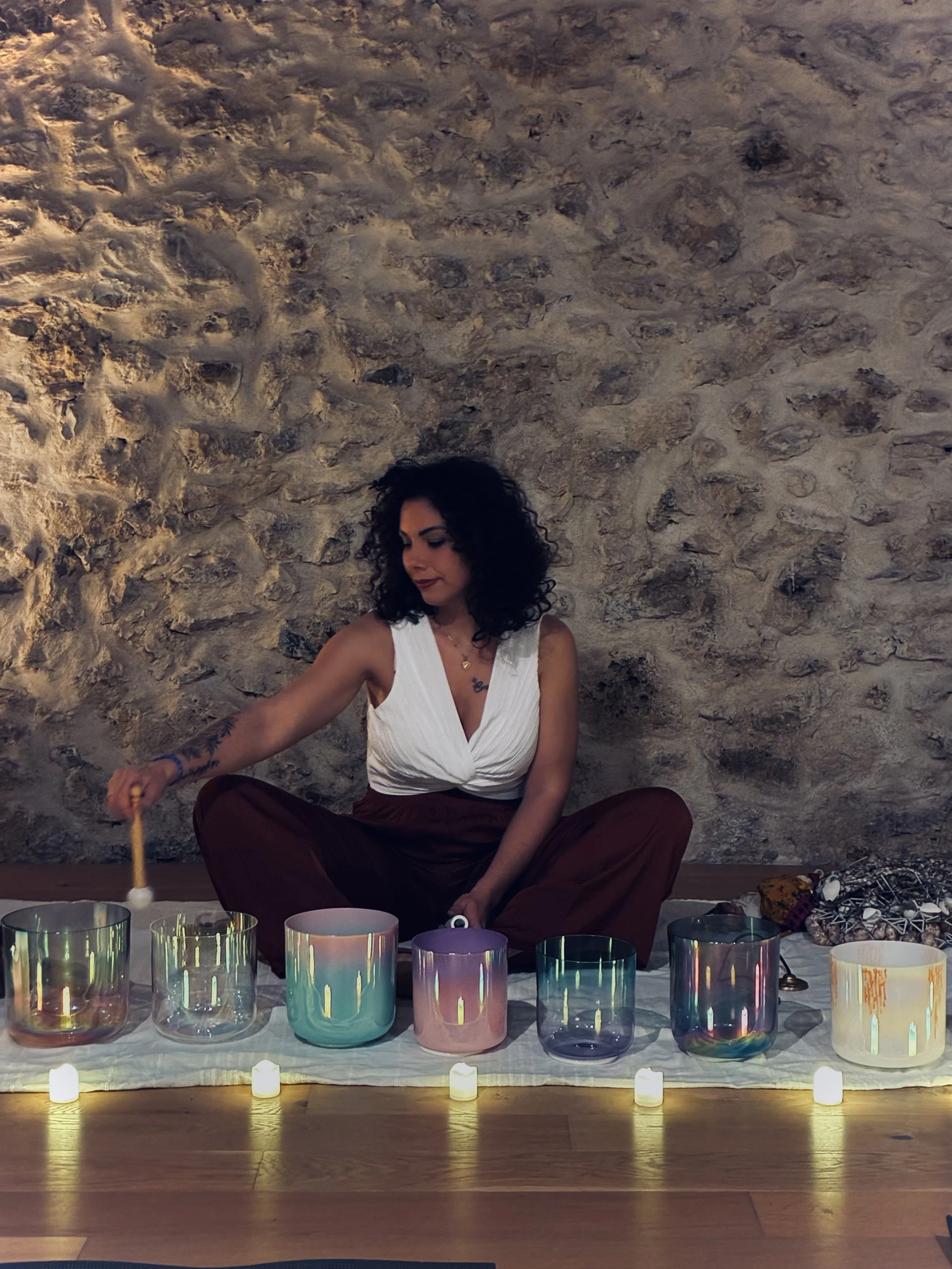 Une femme assise en position de méditation devant un mur de pierre, entourée de plusieurs bougies colorées sur une table. Elle porte un haut blanc et des pantalons marron, avec des tatouages visibles sur ses bras.