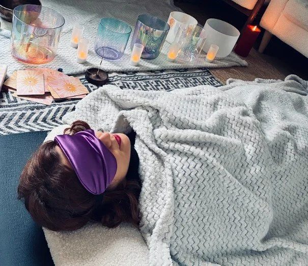 Une femme allongée sur un canapé avec un masque pour dormir violet, entourée de bougies et de cristaux, dans une ambiance relaxante.