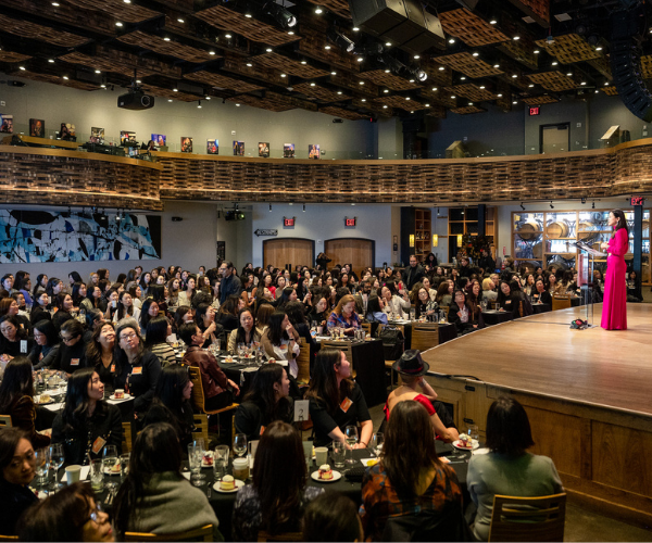 A woman in a pink dress speaking on stage in front of an audience seated at tables in a large, dark auditorium with a balcony and stage lighting.