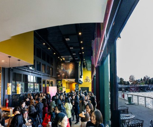 Crowded indoor scene with many people, some wearing masks, at a location with colorful yellow walls, large windows, and a high ceiling with spotlights.