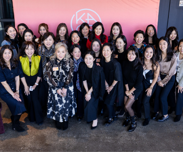 Group of women posing together at a professional event with a pink background and a white circular logo.