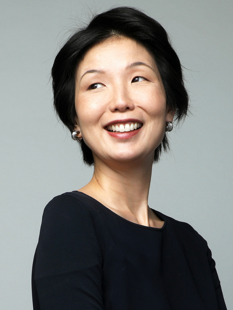 A woman with short black hair smiling and looking to the side, wearing a black top and earrings against a gray background.