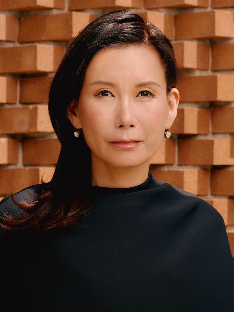 A woman with dark brown hair and fair skin standing in front of a brick wall, wearing a black top and earrings.