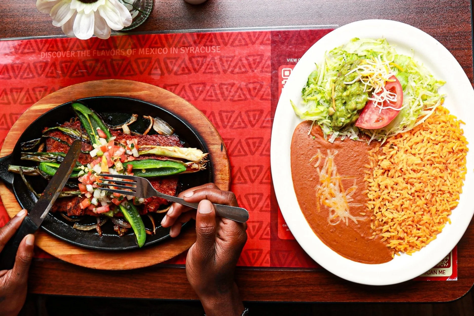 Mexican food on a table includes a sizzling fajita platter with beef, grilled vegetables, and salsa, alongside a plate with refried beans, rice, shredded lettuce, guacamole, and a tomato slice.