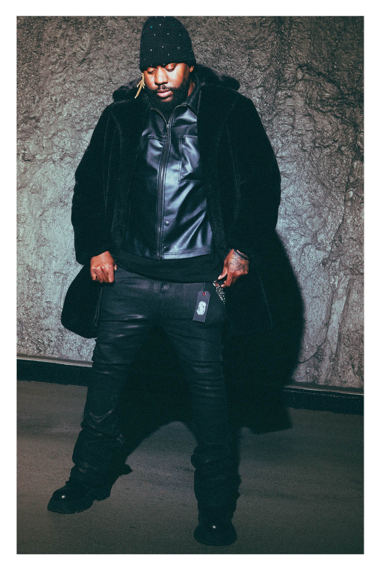 freshmadeit  wearing black leather outfit and knit cap, standing against textured wall.