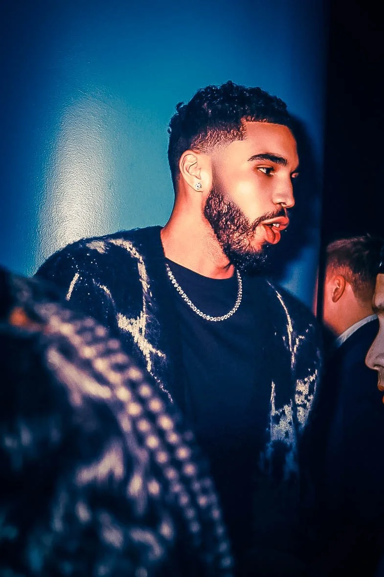 jason Tatum  wearing a black and white jacket and chain necklace, standing against a blue background.