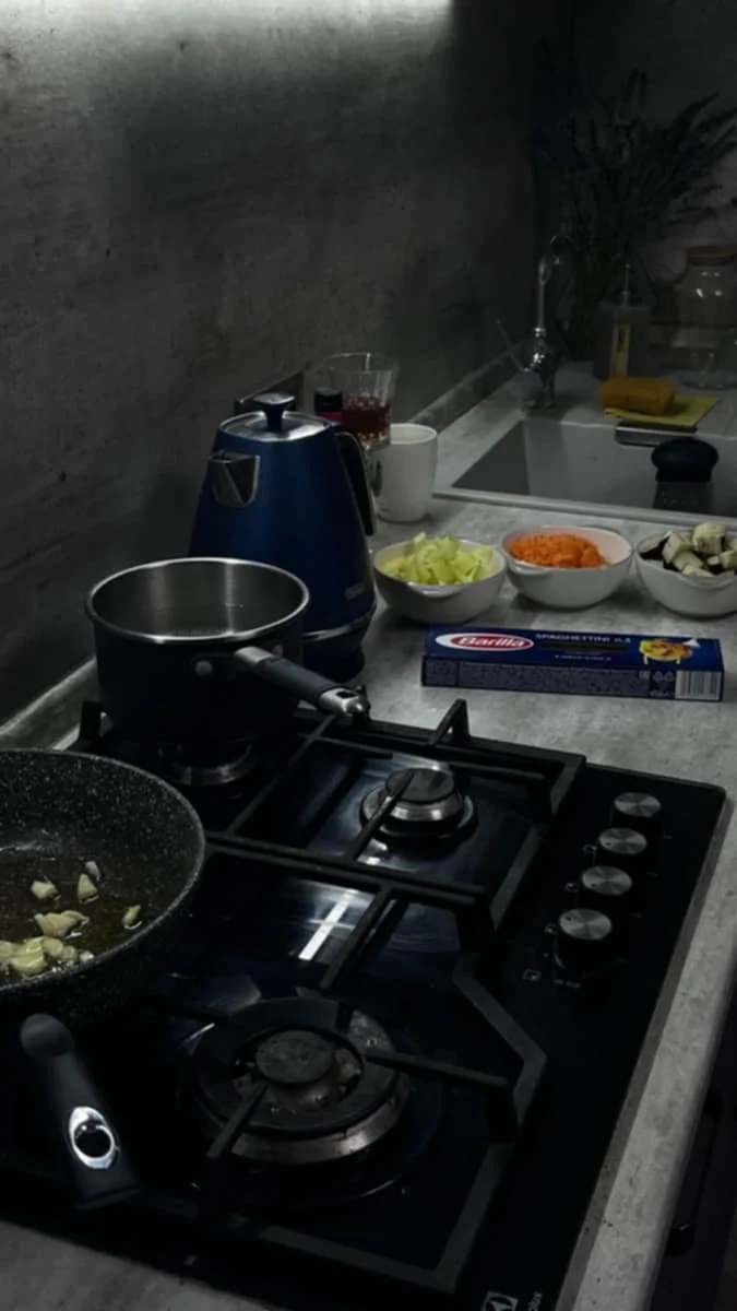 Kitchen countertop with a gas stove, frying pan, boiling pot, several bowls of chopped vegetables, a blue electric kettle, a glass of dark beverage, a white mug, and a sink with a plant in the background.