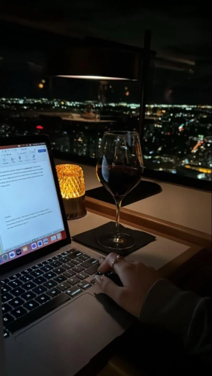 A person working on a laptop at a restaurant table during nighttime, with a glass of red wine, a candle, and a cityscape visible through large windows in the background.