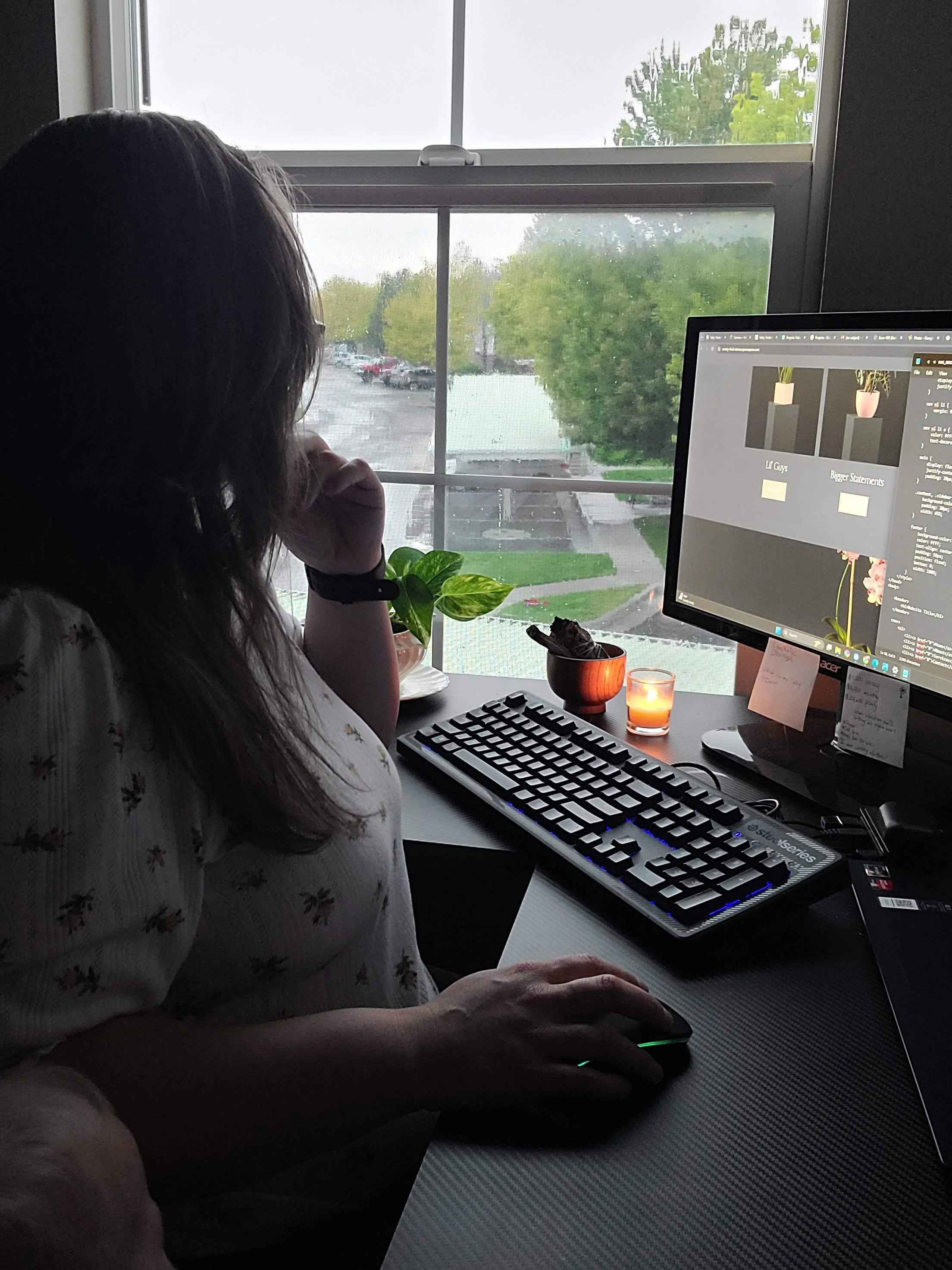 A woman working at her desk with a computer monitor, keyboard, and mouse, viewed from the side, near a window with a view of a parking lot and trees outside.