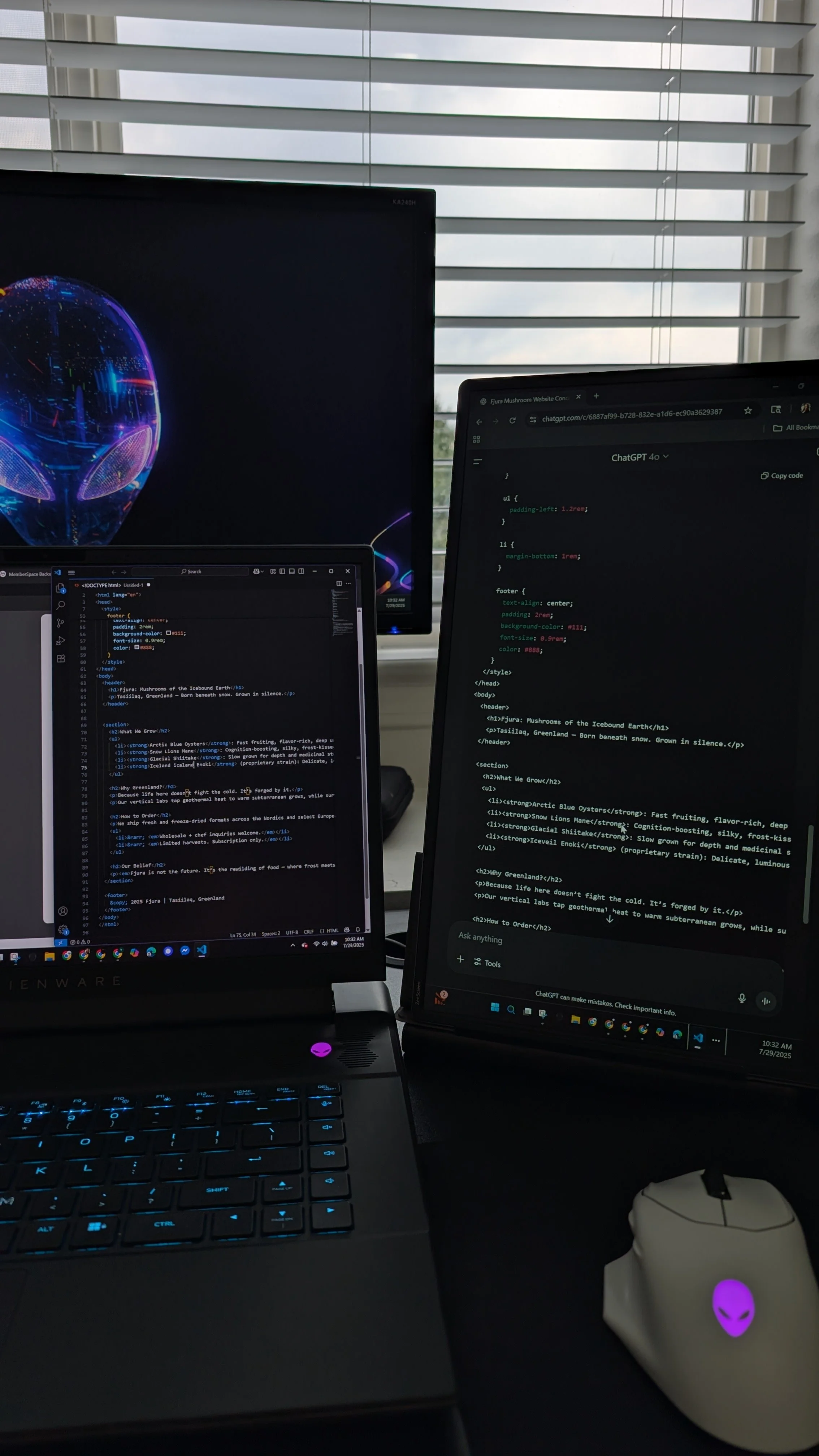 A workspace with a laptop and two monitors displaying code and digital artwork, a computer mouse with an alien face logo, in front of window blinds.