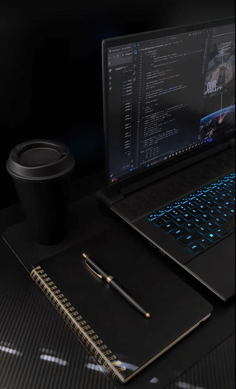 A workspace setup with a laptop displaying code, a black hardcover notebook with a gold and black pen on top, a black disposable coffee cup, and a black mouse pad, all on a dark surface.