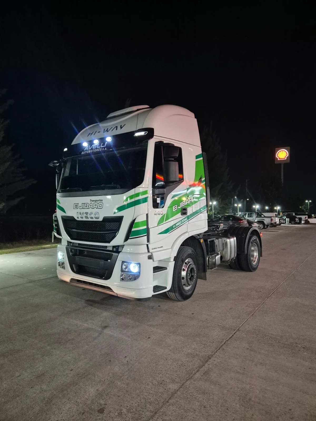 Camión de transporte de carga blanco con detalles verdes en un estacionamiento, noche, con señal de Shell en el fondo.