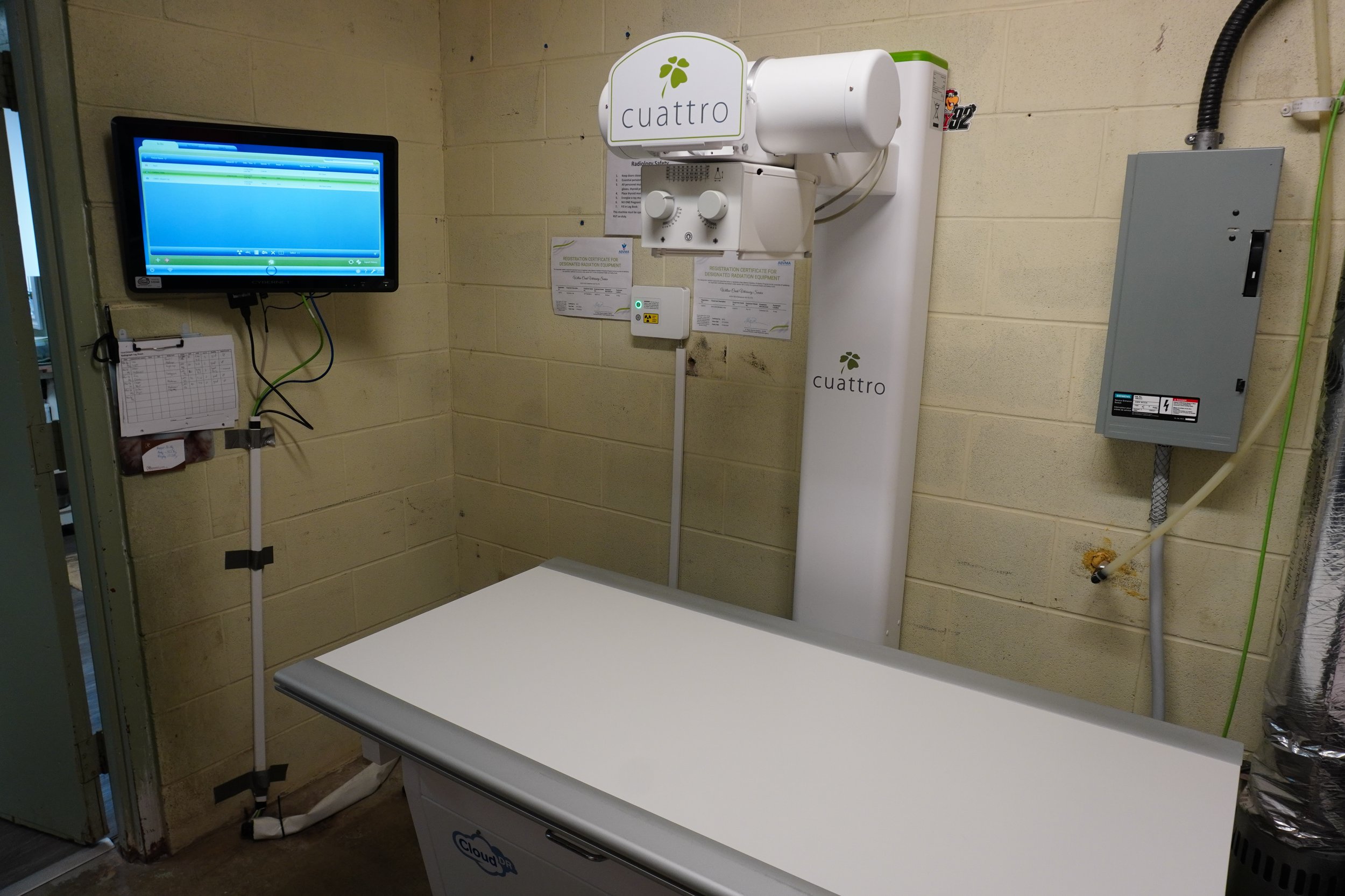 Medical imaging room with a white medical scan table, a computer monitor on the wall, and radiation equipment labeled 'cuattro' on the wall.