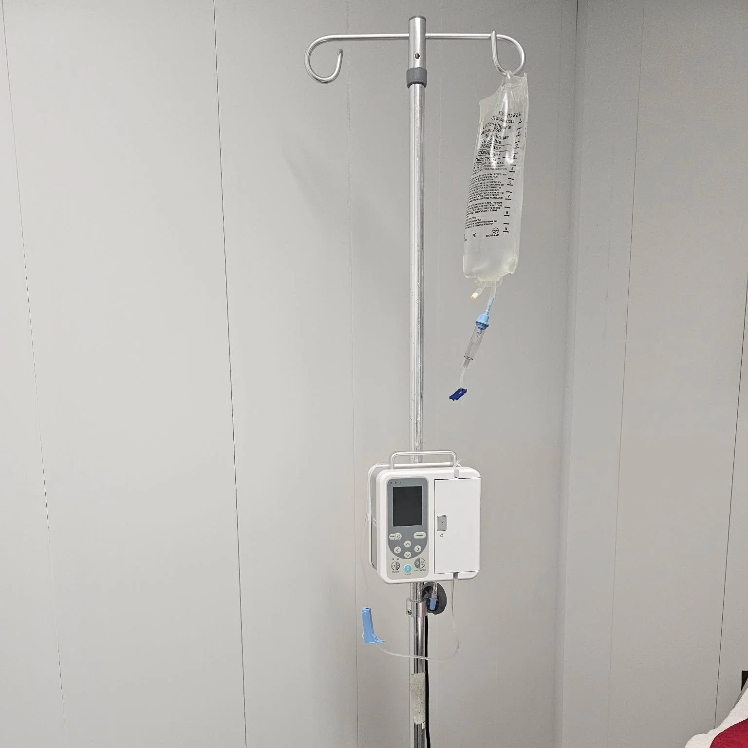 Hospital IV stand with a connected IV bag and infusion pump against a plain white wall.