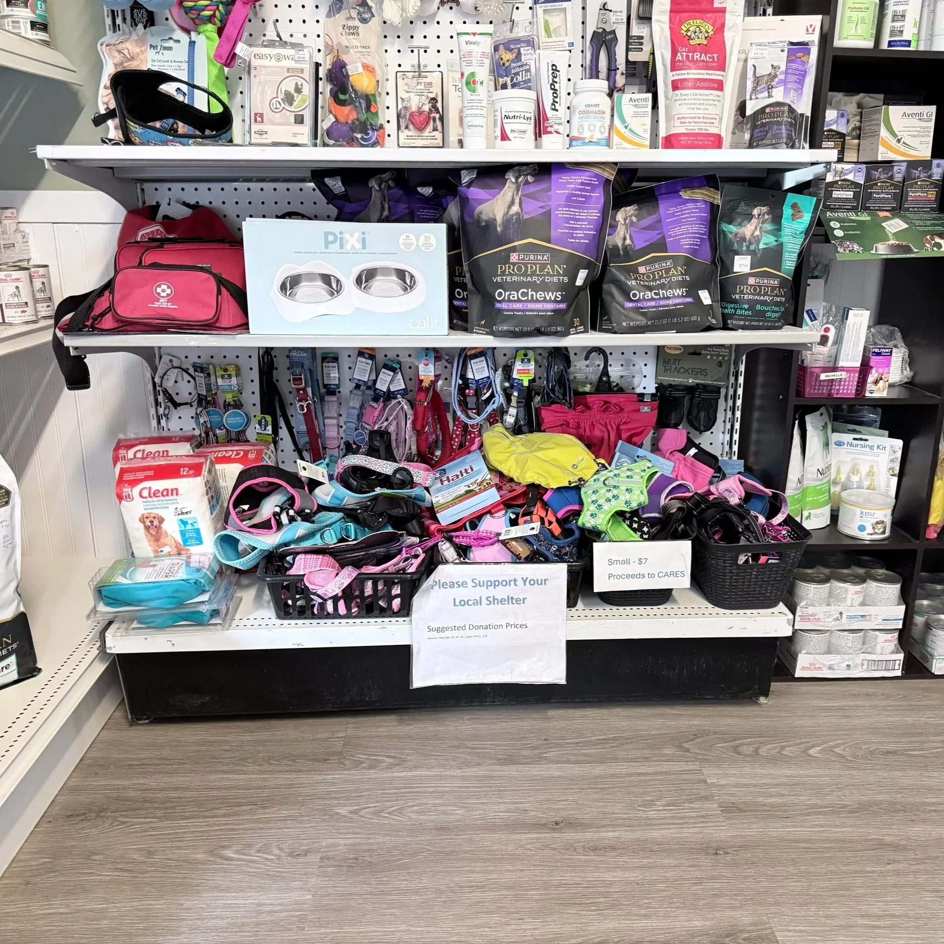 Display of pet supplies, including harnesses, collars, and a variety of pet food in a pet store. Sign indicating donation proceeds for pet shelter.