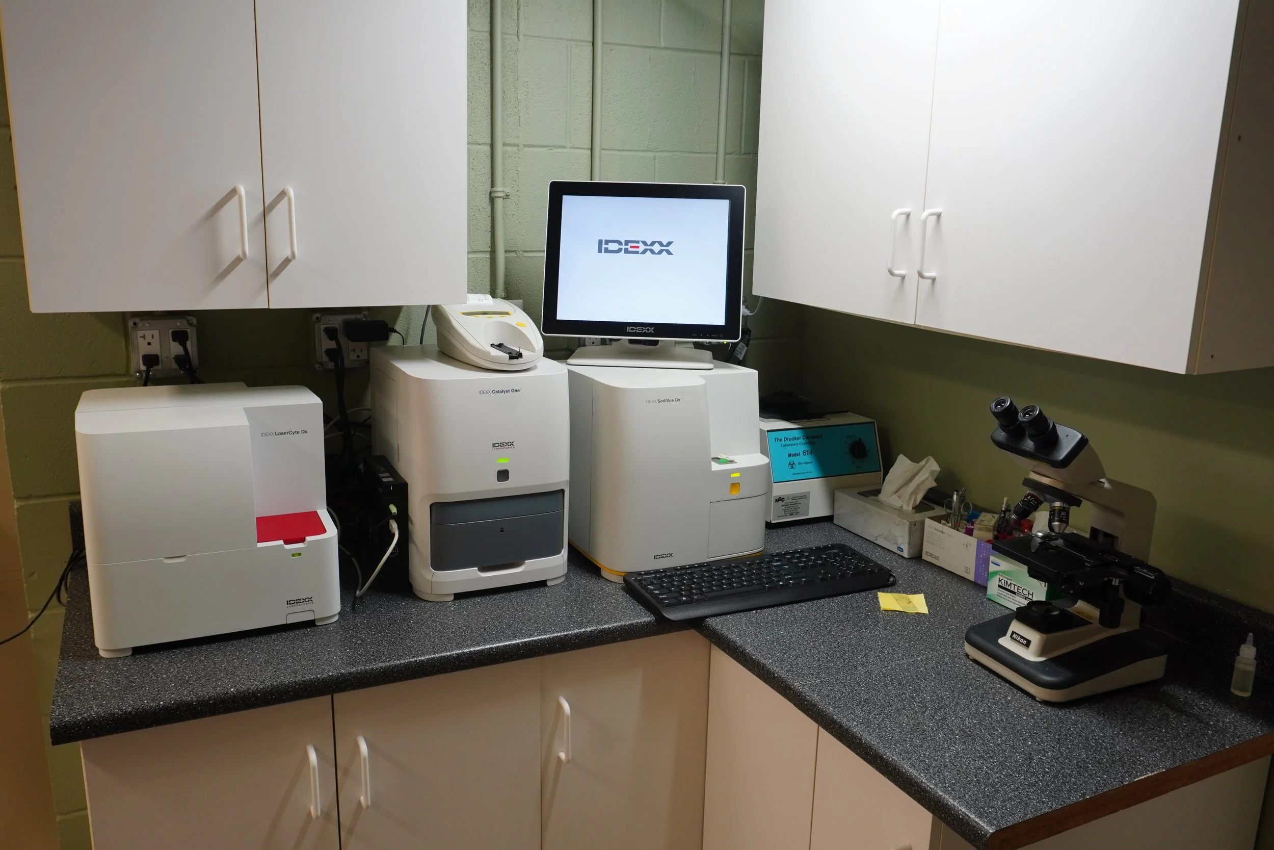 Medical laboratory workstation with equipment including DNA analysis machines, a computer monitor, a tissue culture microscope, and supplies on a black countertop.