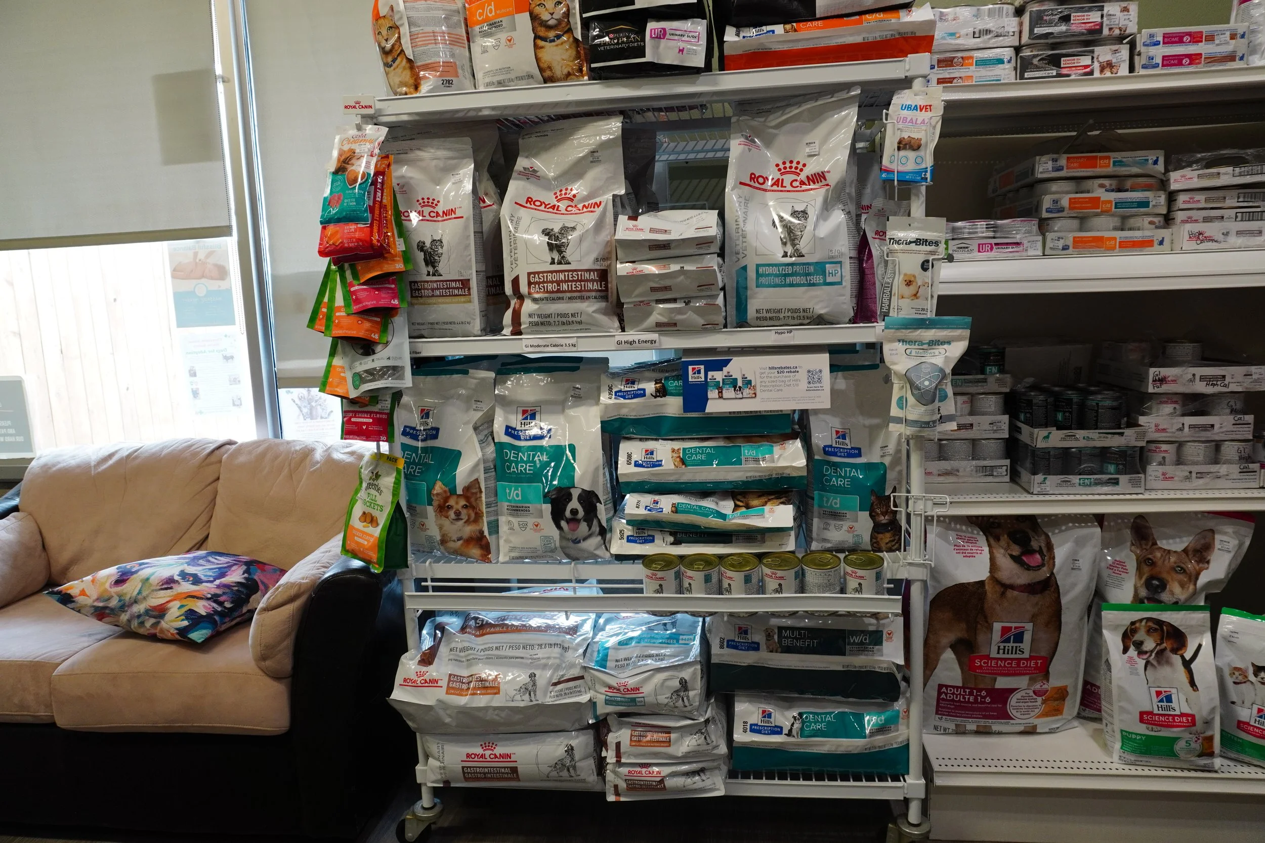 Shelves of pet food and supplies in a pet store, with bags of dog and cat food displayed, and a beige couch with a colorful pillow on the left side.