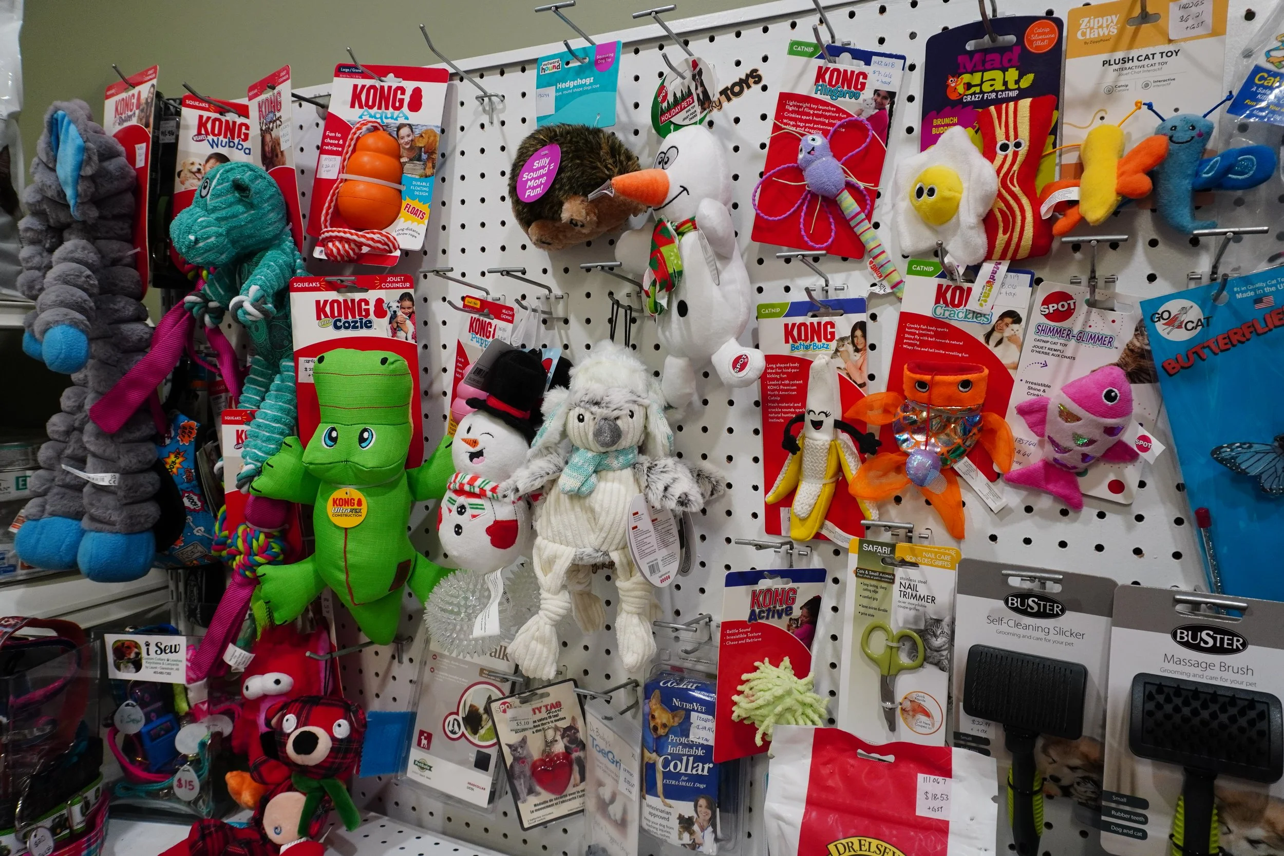 A display of various pet toys and accessories on a white pegboard. Includes plush toys, squeakers, and grooming tools.