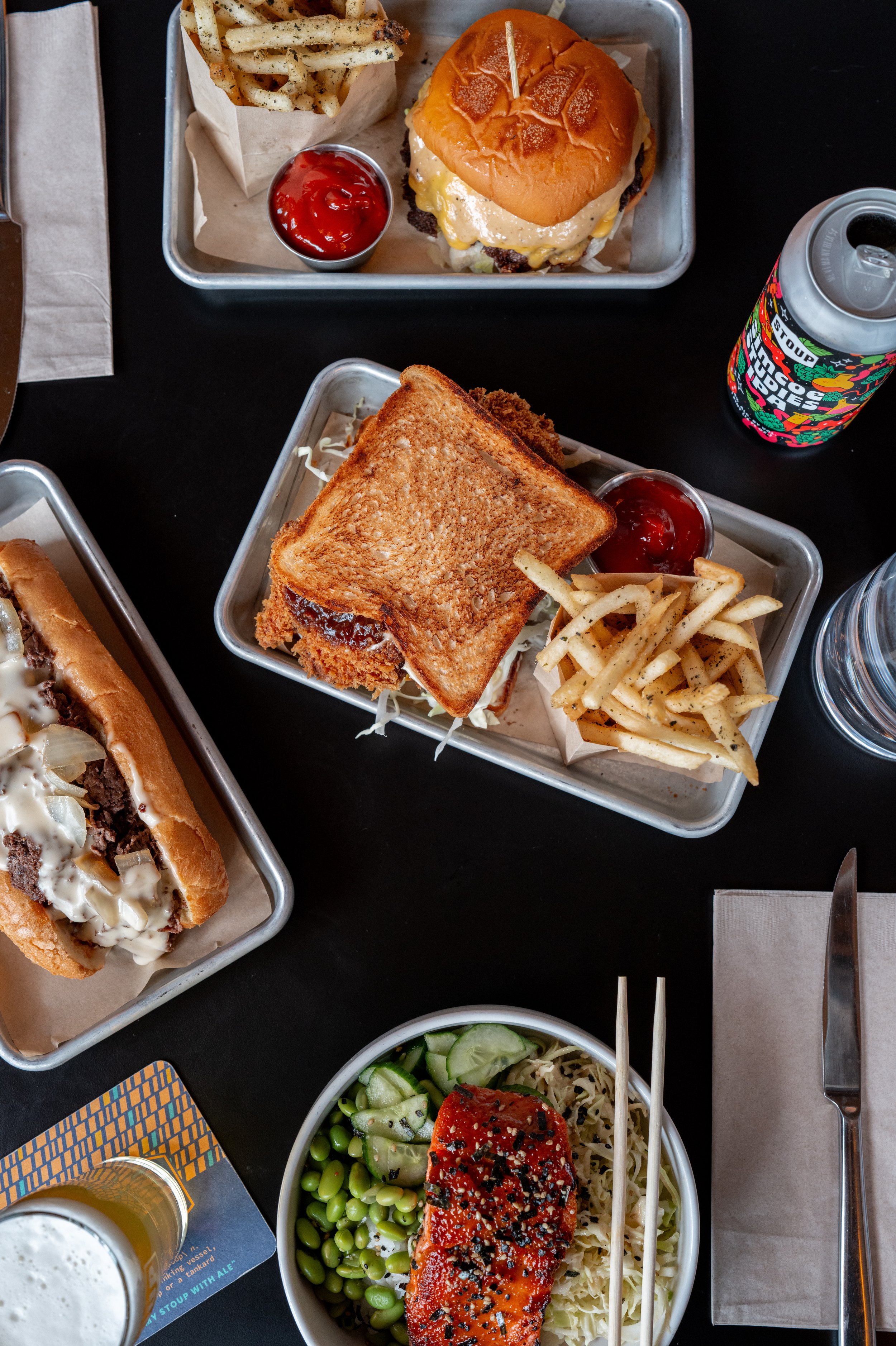 Top view of a meal on black table, including cheeseburger with fries and ketchup, toasted sandwich with fries and ketchup, salad with salmon, cucumber, and edamame, and a can of soda.