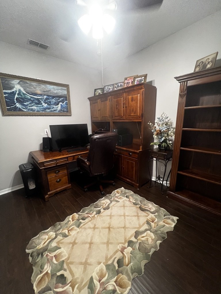 Home office with wooden desk and hutch, computer monitor, leather chair, area rug, bookshelf, framed painting, and decorative flowers.