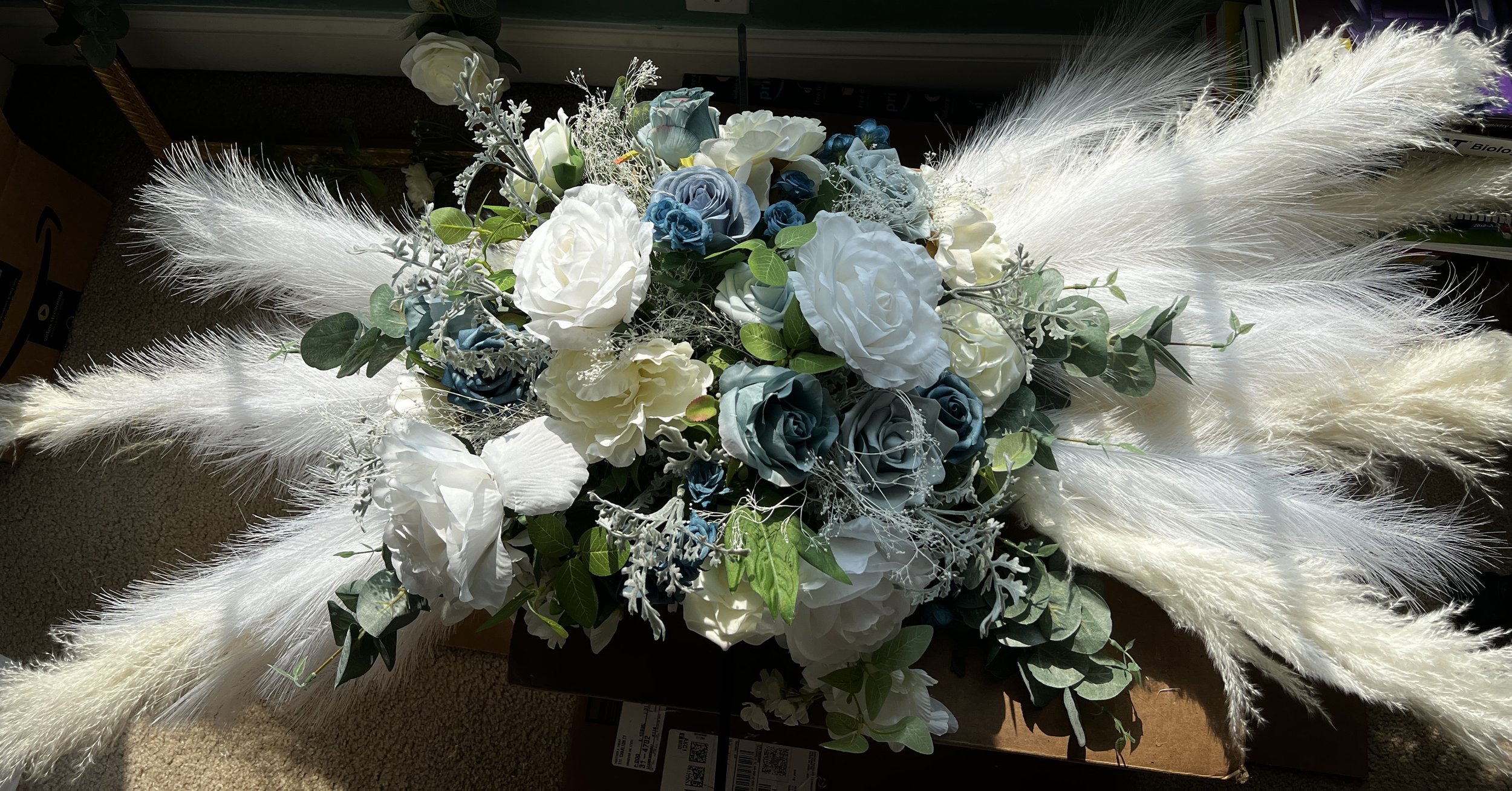 A floral arrangement featuring white and blue artificial roses, pampas grass, and greenery.