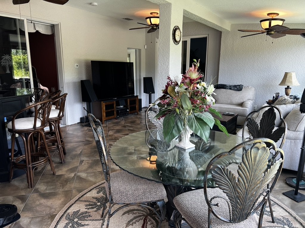 A living room with a glass dining table and ornate chairs, featuring a floral centerpiece. There's a large flat-screen TV positioned on a stand. The room includes comfortable seating, bar stools, ceiling fans, and a decorative rug on a tiled floor.