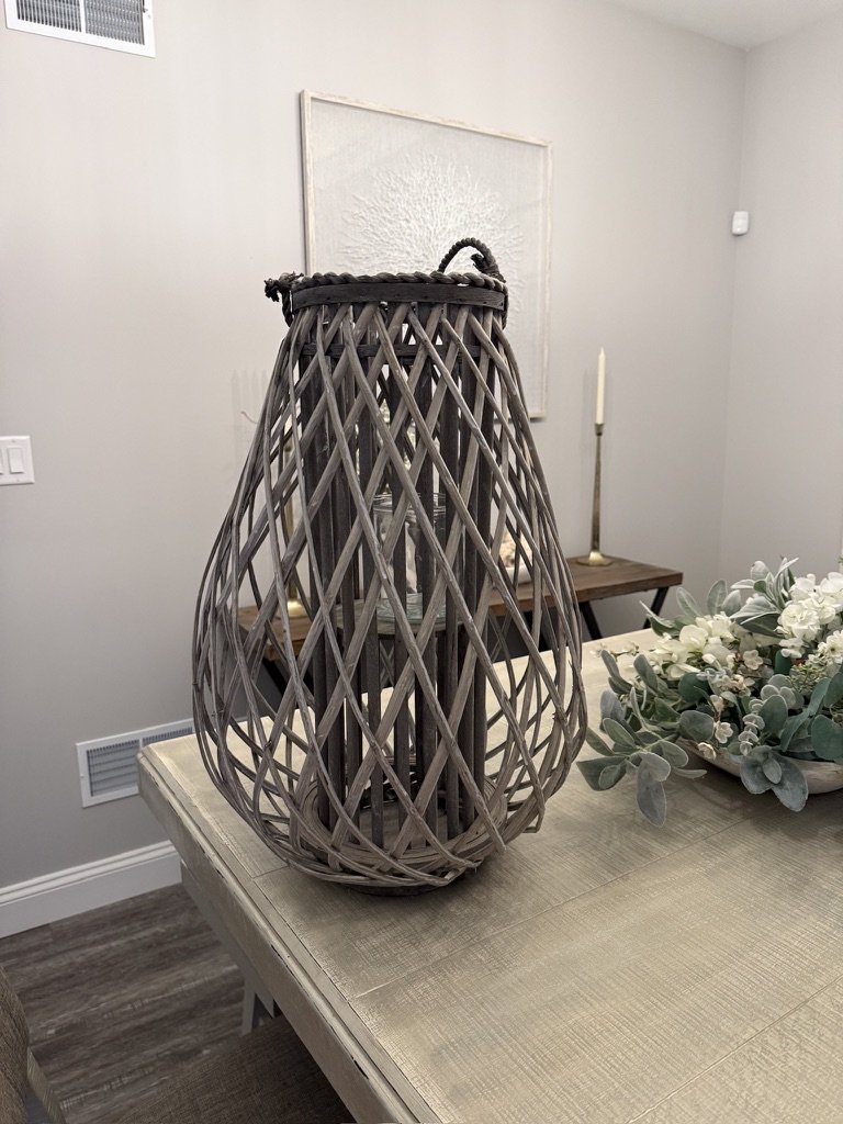 Wicker lantern with glass candle holder on a wooden table, next to a floral centerpiece with white flowers and greenery, in a dining room with neutral decor.
