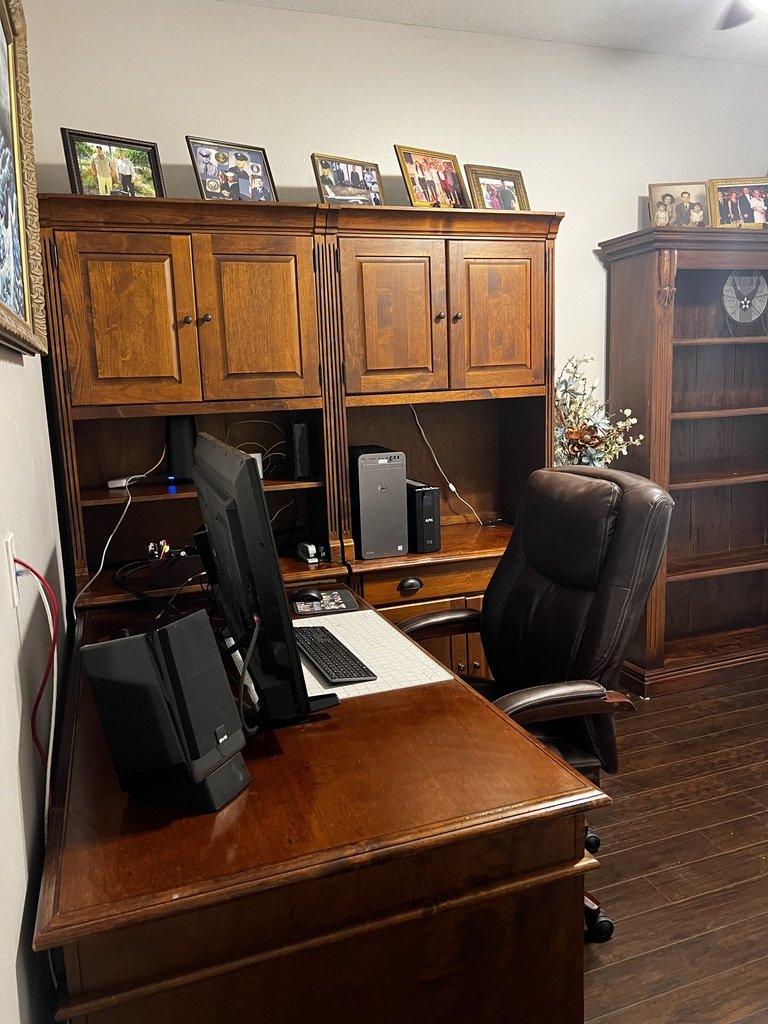 Home office with a wooden desk, computer setup, and leather chair. Decorative cabinets on the desk hold family photos. An empty wooden bookshelf is in the background.