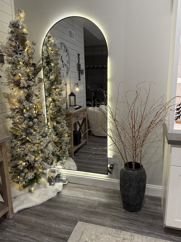 Interior with LED mirror, decorative Christmas tree, vase with branches, wall clock, lantern, and wooden flooring.