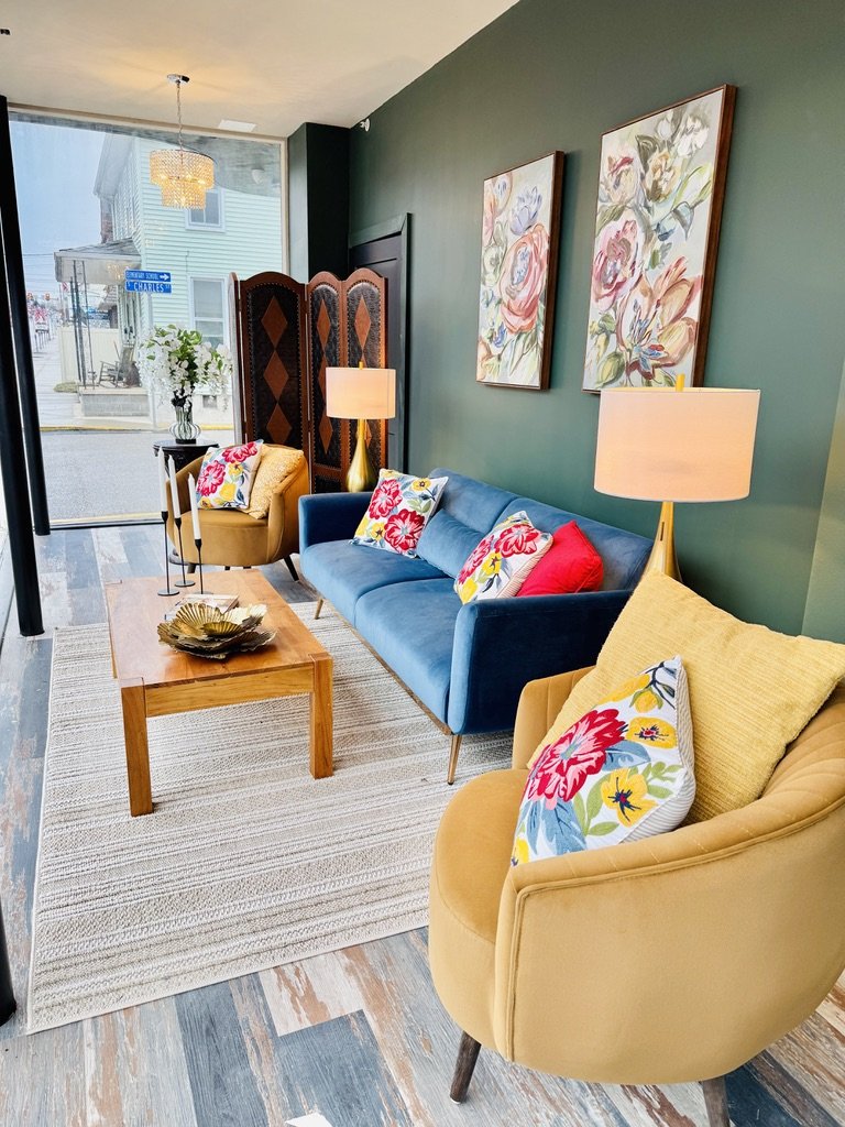 Cozy living room with dark green walls, featuring a blue sofa and yellow armchairs adorned with colorful floral pillows. A wooden coffee table and lamps brighten the space. Artwork and a decorative screen add character. Large window allows natural li