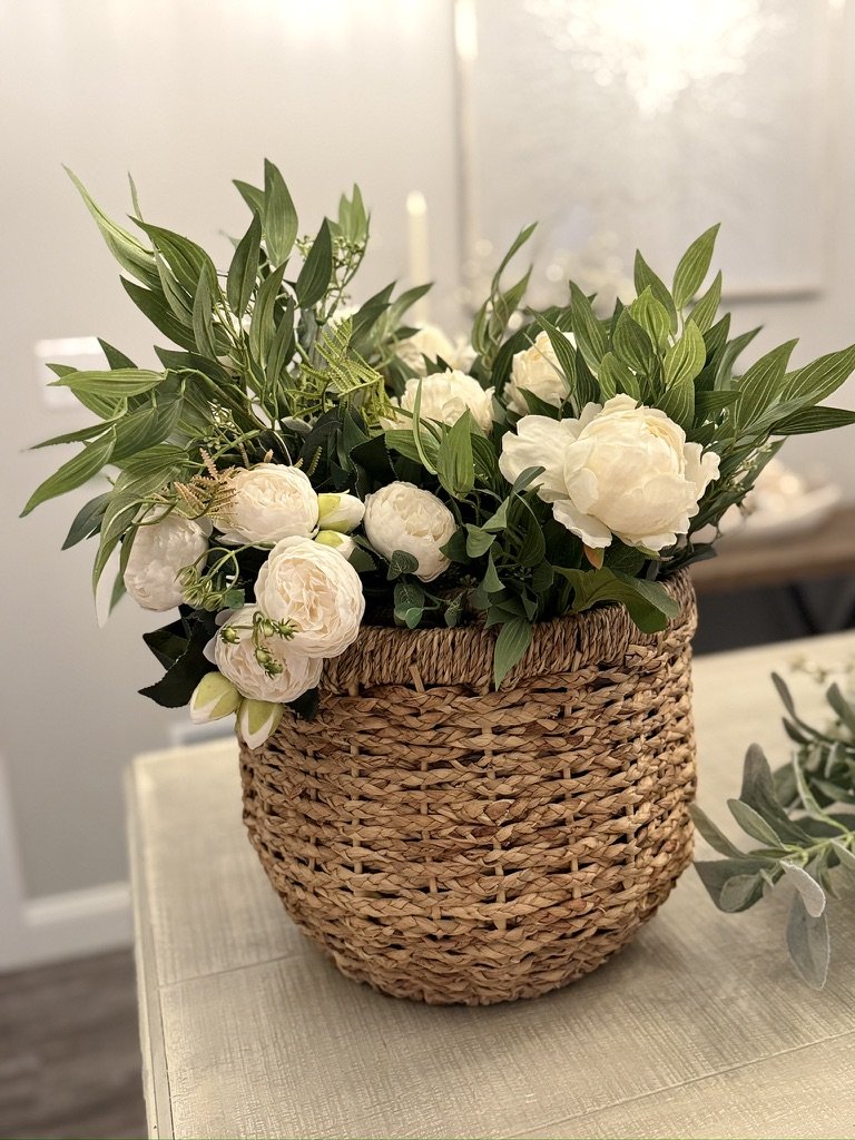 White peonies and green leaves in a wicker basket on a table against a neutral background.