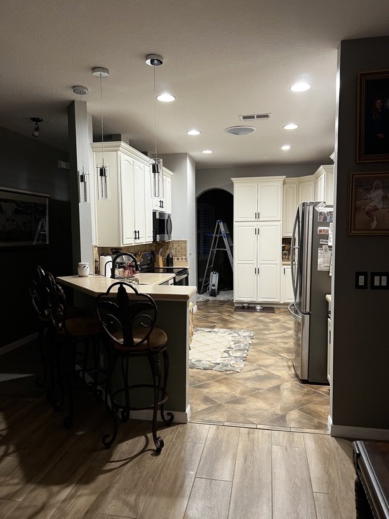 Modern kitchen with white cabinets, stainless steel appliances, bar stools at a counter, pendant lights, and dark flooring.