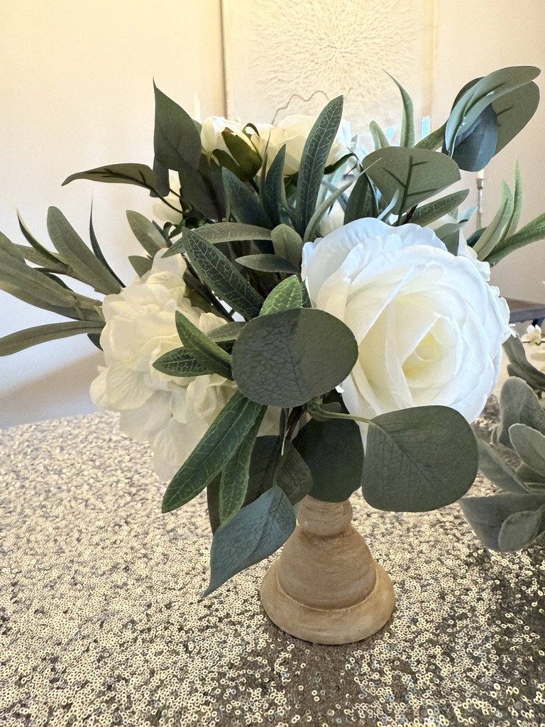 Artificial white rose and greenery arrangement in a wooden vase on a sparkly silver tablecloth.