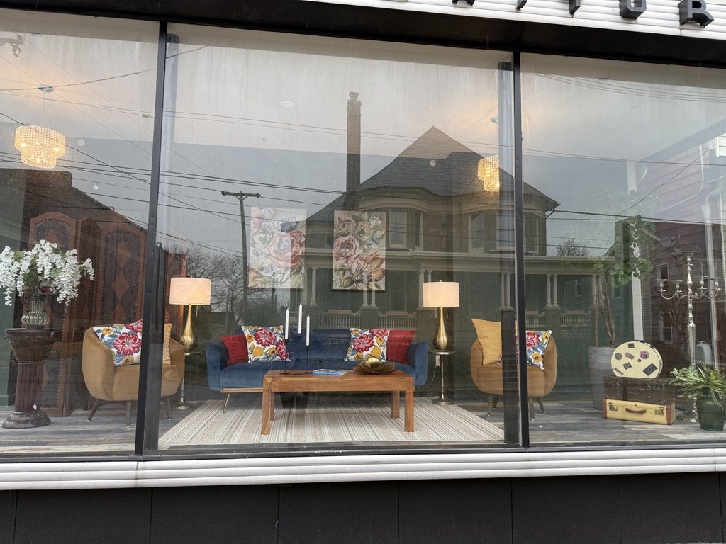 Store window display showing living room furniture, including a blue sofa with colorful floral pillows, a wooden coffee table, floor lamps, and decorative art pieces. A floral arrangement, folding screen, and suitcases are also visible.