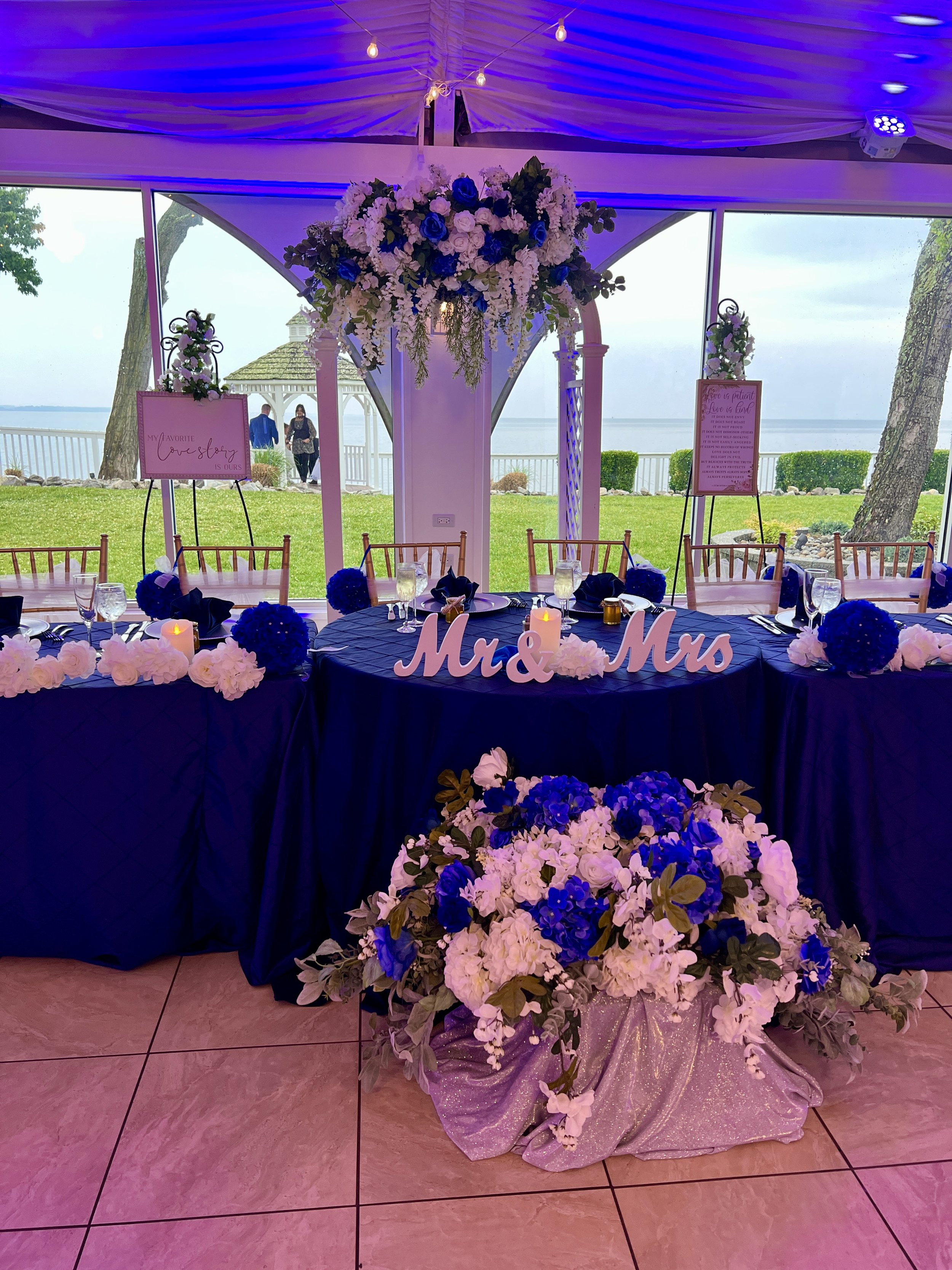 Wedding reception table with blue and white decor, including a large floral centerpiece, "Mr & Mrs" sign, and outdoor lake view.