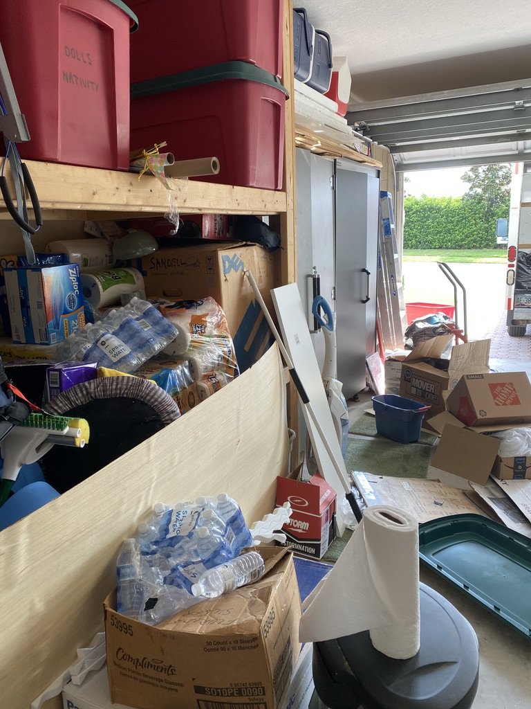 Cluttered garage with storage bins, cardboard boxes, water bottles, and miscellaneous items.