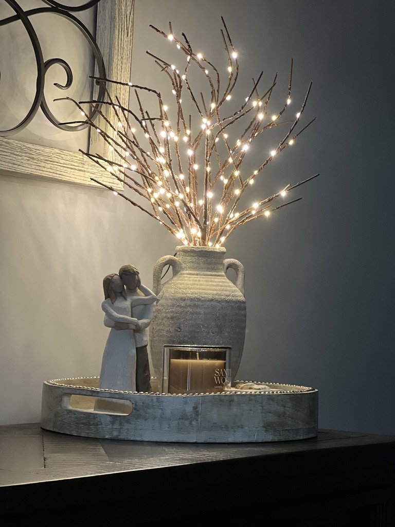 A decorative setup featuring a rustic vase filled with twinkling branch lights, a small figurine of a couple embracing, and a scented candle on a round wooden tray.