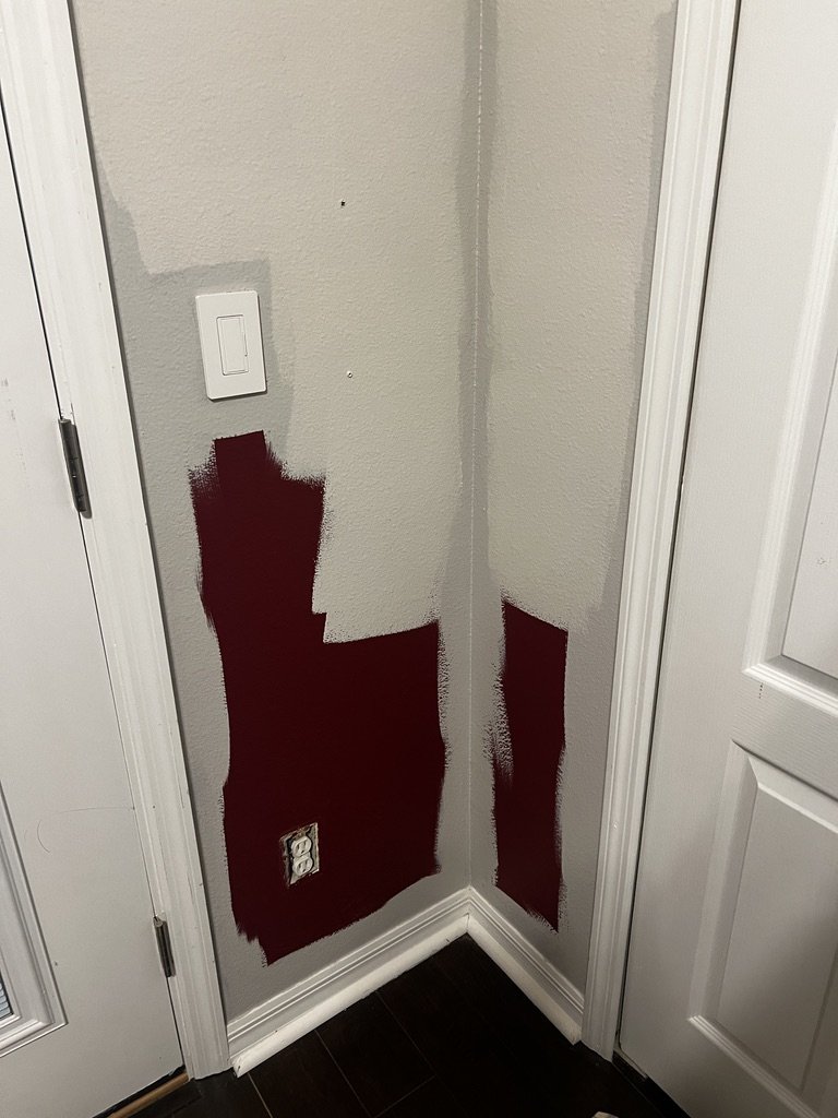 A corner interior wall with partial maroon paint patches over a light gray base, near a white door and light switch.