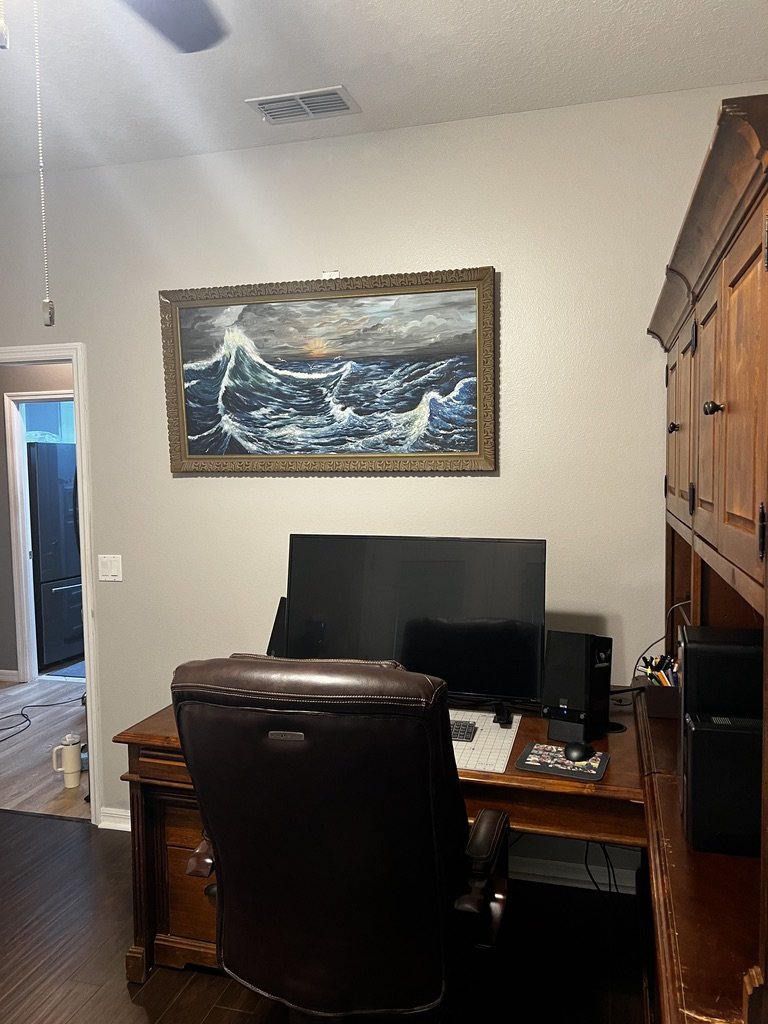 Home office setup with a large computer monitor on a wooden desk, a leather office chair, and a framed ocean waves painting on the wall.
