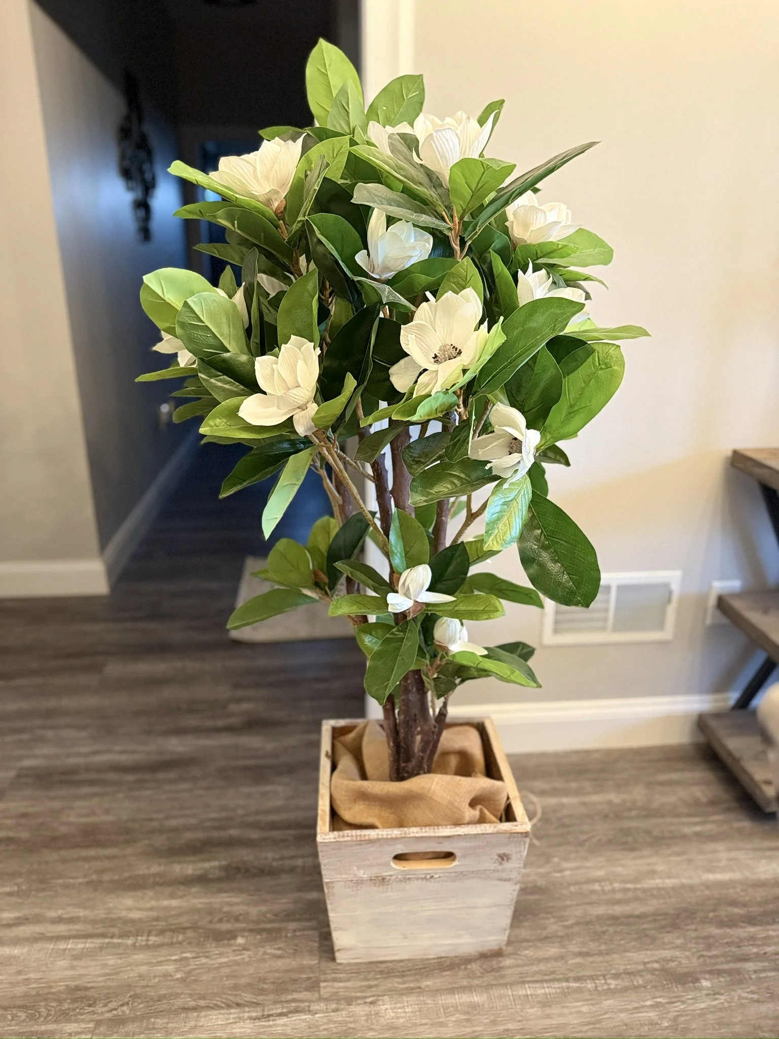 Decorative artificial plant with white flowers in a wooden box on a wooden floor.