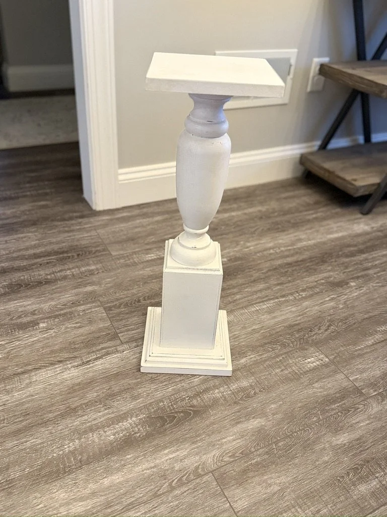 Small white decorative pedestal with square top and base, standing on a hardwood floor.