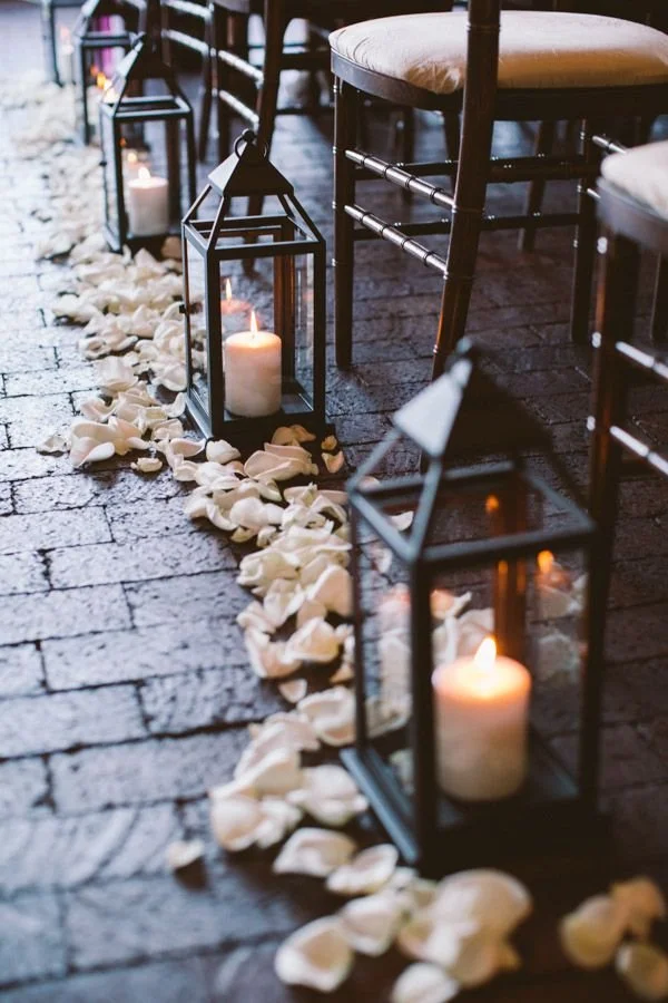 Pathway decorated with white rose petals and black lanterns with lit candles beside rows of chairs.