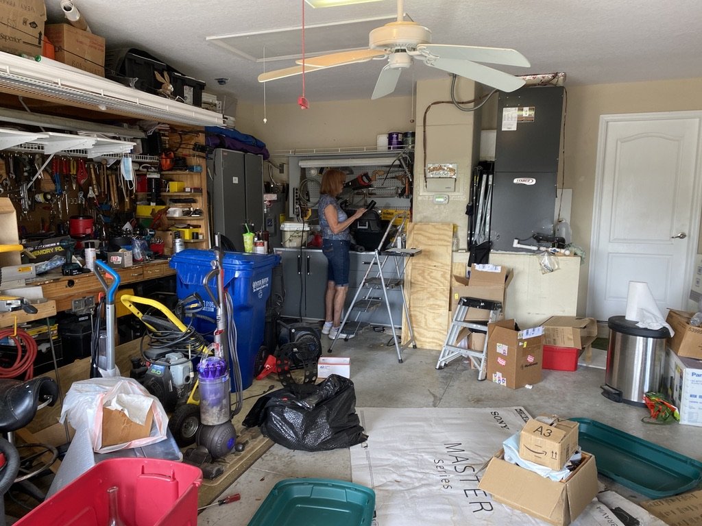 Cluttered garage with a person organizing items; visible items include storage shelves, recycling bins, tools, boxes, and a ceiling fan.