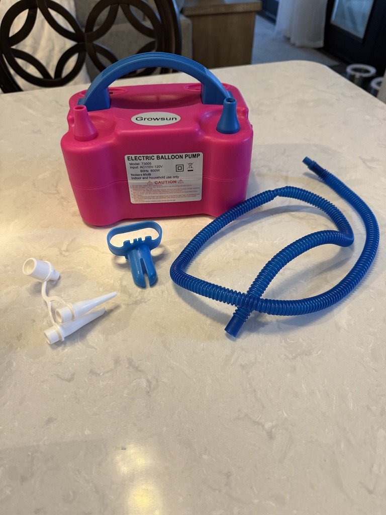 Pink electric balloon pump with blue hose and nozzles on a light-colored surface.