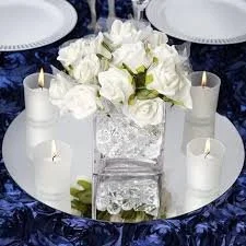 Table centerpiece with white roses in a glass vase, surrounded by lit candles and placed on a mirrored surface.