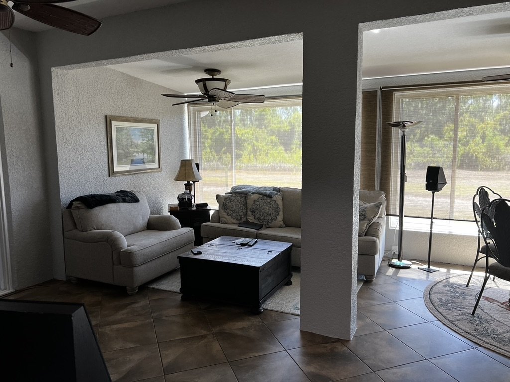 Living room with beige sofas, a wooden coffee table, and large windows. The room has ceiling fans, a floor lamp, and tiled flooring. There is a round rug and a framed picture on the wall.