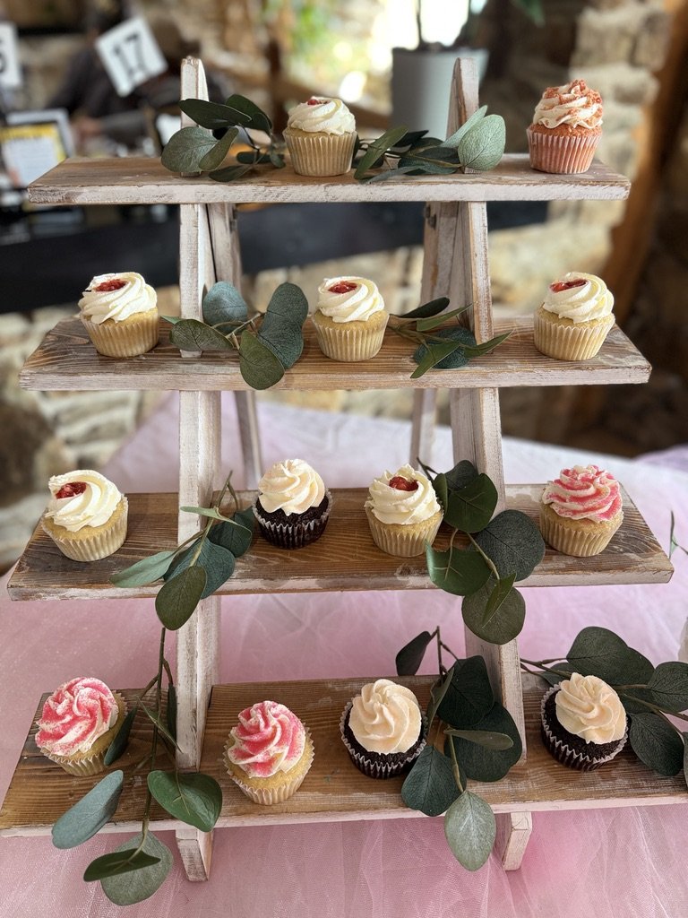 Cupcakes on a tiered wooden stand decorated with greenery on a pink cloth background.