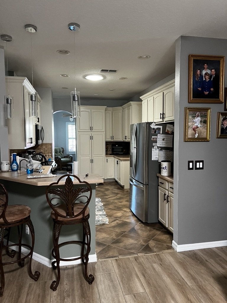 Modern kitchen with white cabinets, stainless steel appliances, pendant lights, bar stools, and family photos on the wall.