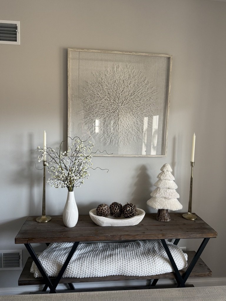 Minimalist home decor setup with a wooden console table featuring a white vase with branches, a decorative white tree, pinecones in a bowl, and two tall candles, under a framed art piece.