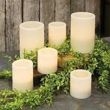 Six ivory pillar candles surrounded by green foliage on a rustic wooden surface.